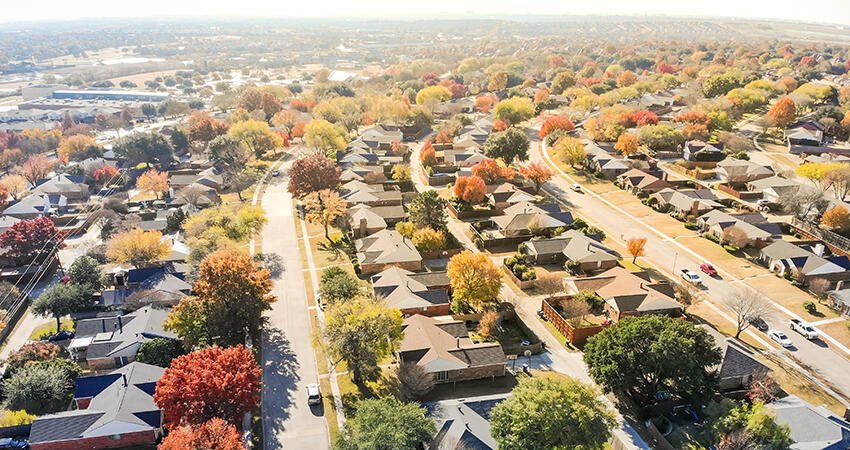 Aerial view of a suburban residential neighborhood with rows of single-family houses surrounded by autumn foliage and tree-lined streets, showing rooftops, driveways, and yards from above.
