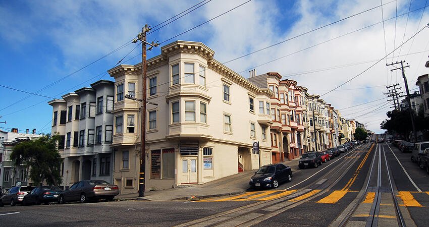 A steep city street lined with multi-story apartment buildings, parked cars, and cable car tracks running uphill under a partly cloudy sky