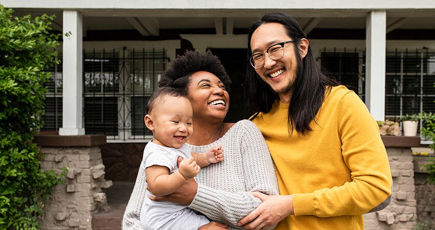 Two adults smiling and holding a laughing baby while standing together on the front porch of a home