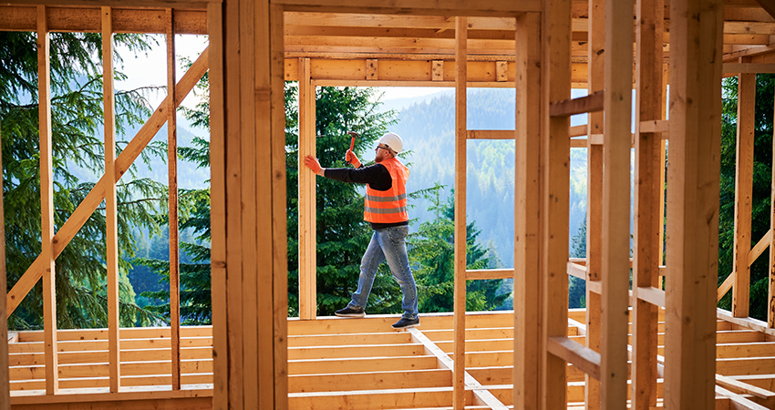 Carpenter constructing two-story wooden frame house near forest. Man hammering nails into the structure, wearing protective helmet and construction vest. Concept of modern ecological construction.