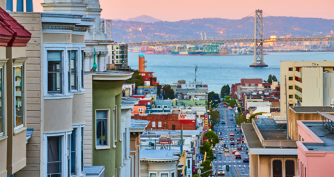 Colorful Victorian-style houses line a steep street in San Francisco, leading down toward the bay at sunset, with the Bay Bridge visible in the distance and city lights beginning to glow.