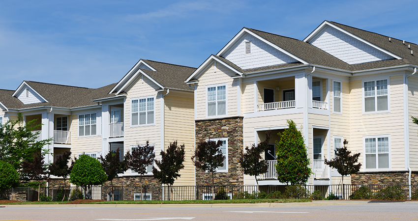 A row of modern two-story apartment buildings with beige siding, white trim, and stone accents, featuring balconies and large windows, lined with small trees and a sidewalk along the street under a blue sky.
