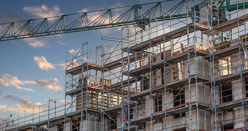 Multi-story building under construction surrounded by metal scaffolding, with a tall green crane overhead against a partly cloudy sky at sunset.