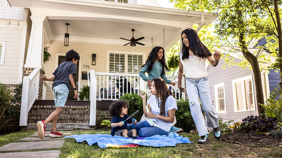 People gather outside a house while someone blows bubbles for a child on a sunny day.