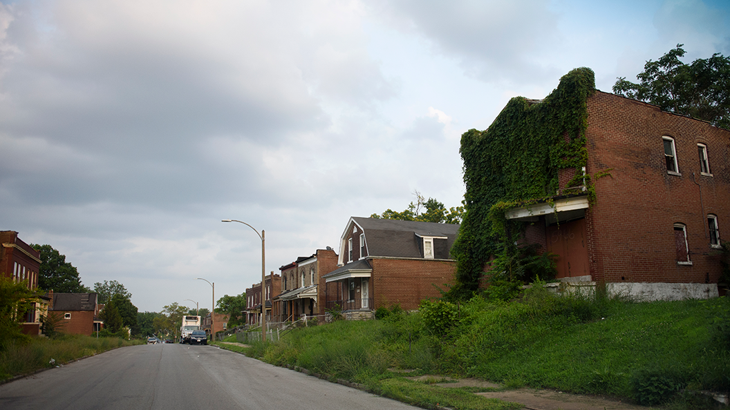 Overgrown ivy-covered brick house on a quiet street with other aging homes under a cloudy sky.