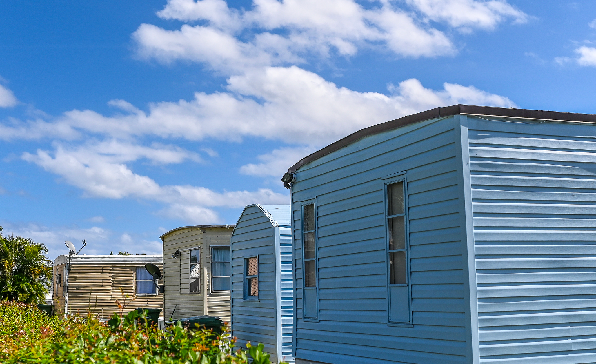 Row of light-colored mobile homes or manufactured houses under a bright blue sky with scattered white clouds.