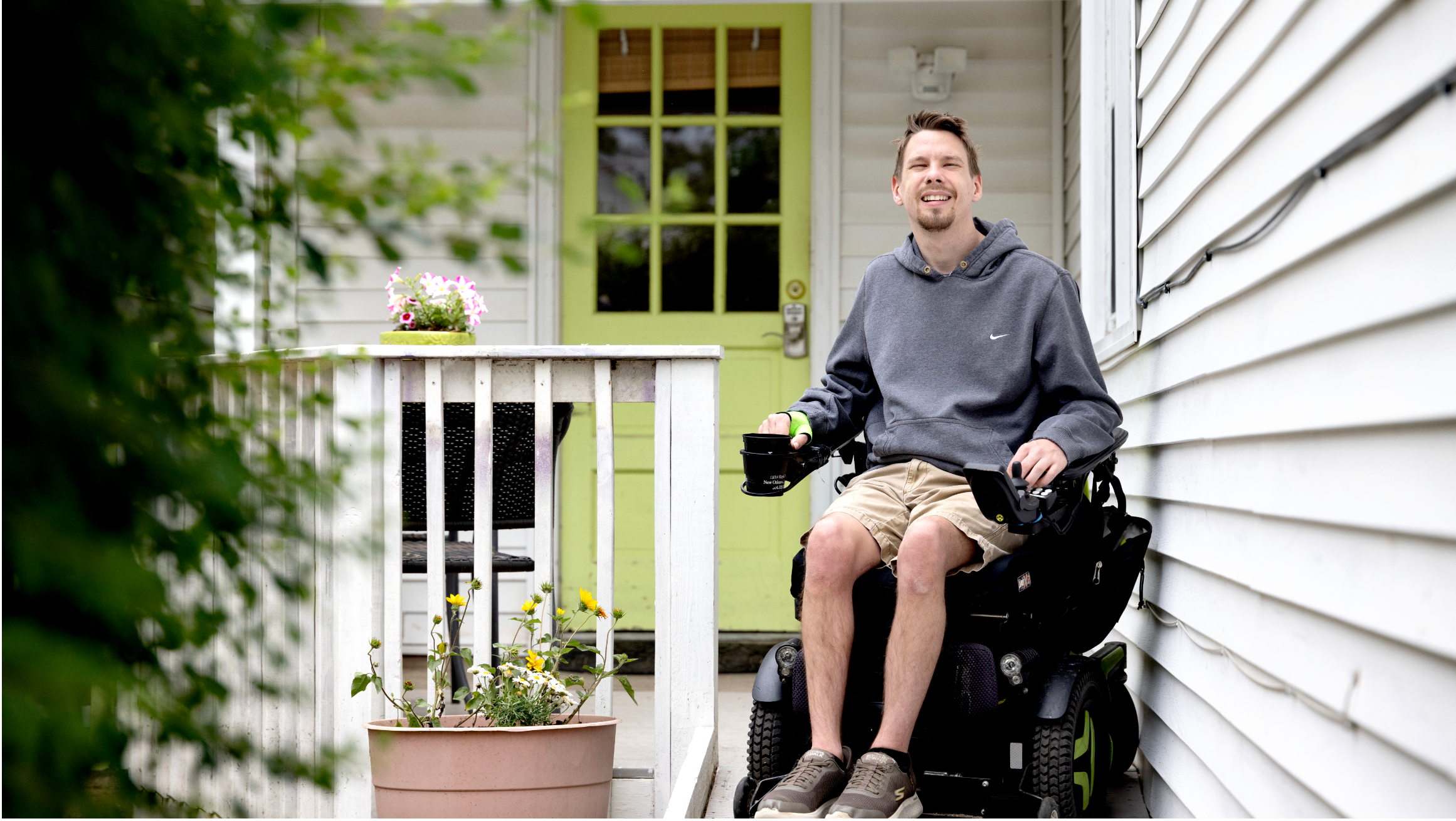 Person using a powered wheelchair on a home porch ramp beside a light green front door and potted flowers.