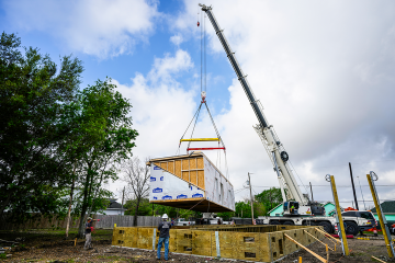 Workers guide a crane‑lifted modular home section onto its foundation at a construction site