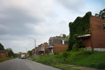 Overgrown ivy-covered brick house on a quiet street with other aging homes under a cloudy sky.