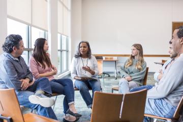 A small group of people seated in a circle during a discussion in a bright meeting room with large windows.