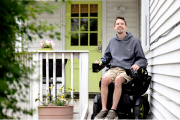 Person using a powered wheelchair on a home porch ramp beside a light green front door and potted flowers.