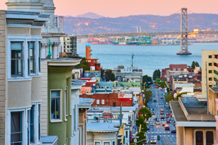 Colorful Victorian-style houses line a steep street in San Francisco, leading down toward the bay at sunset, with the Bay Bridge visible in the distance and city lights beginning to glow.