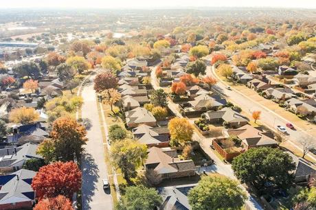 Aerial view of a suburban residential neighborhood with rows of single-family houses surrounded by autumn foliage and tree-lined streets, showing rooftops, driveways, and yards from above.