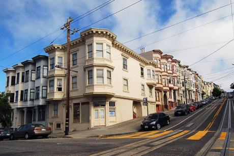 A steep city street lined with multi-story apartment buildings, parked cars, and cable car tracks running uphill under a partly cloudy sky