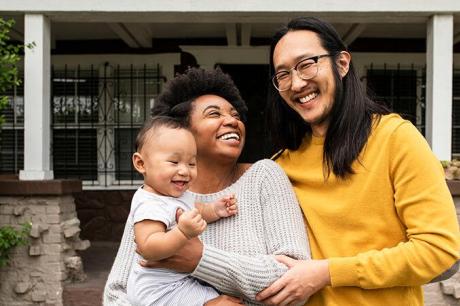 Two adults smiling and holding a laughing baby while standing together on the front porch of a home