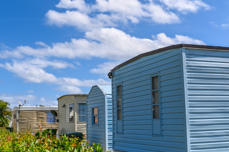 Row of light-colored mobile homes or manufactured houses under a bright blue sky with scattered white clouds.