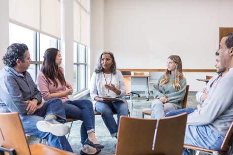 A small group of people seated in a circle during a discussion in a bright meeting room with large windows.