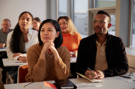 Group of adults seated at desks in a classroom or workshop, listening attentively and taking notes in a bright meeting room.