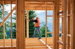 Carpenter constructing two-story wooden frame house near forest. Man hammering nails into the structure, wearing protective helmet and construction vest. Concept of modern ecological construction.