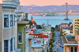Colorful Victorian-style houses line a steep street in San Francisco, leading down toward the bay at sunset, with the Bay Bridge visible in the distance and city lights beginning to glow.