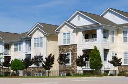 A row of modern two-story apartment buildings with beige siding, white trim, and stone accents, featuring balconies and large windows, lined with small trees and a sidewalk along the street under a blue sky.