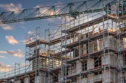 Multi-story building under construction surrounded by metal scaffolding, with a tall green crane overhead against a partly cloudy sky at sunset.